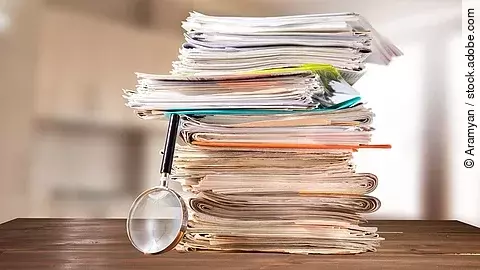 Dokumente Auf einem Tisch liegt ein Stapel mit Unterlagen und daneben eine Lupe.