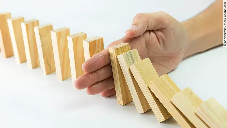 Hölzerne Dominosteine sind in einer Reihe aufgebaut. Einige davon sind bereits umgefallen, eine Hand stoppt den Dominoeffekt.