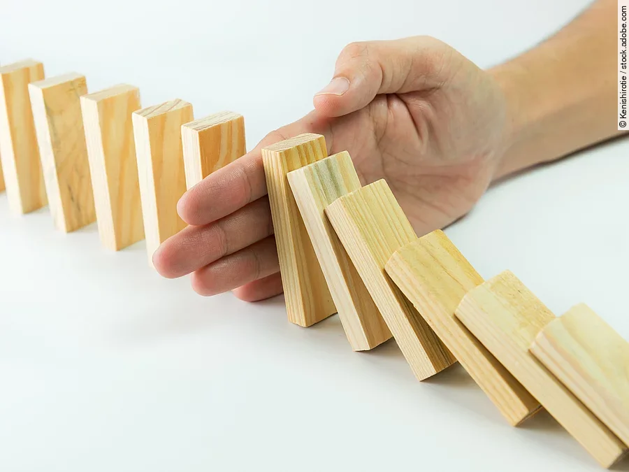Hölzerne Dominosteine sind in einer Reihe aufgebaut. Einige davon sind bereits umgefallen, eine Hand stoppt den Dominoeffekt.