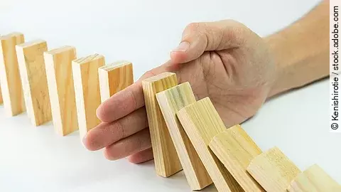Hölzerne Dominosteine sind in einer Reihe aufgebaut. Einige davon sind bereits umgefallen, eine Hand stoppt den Dominoeffekt.