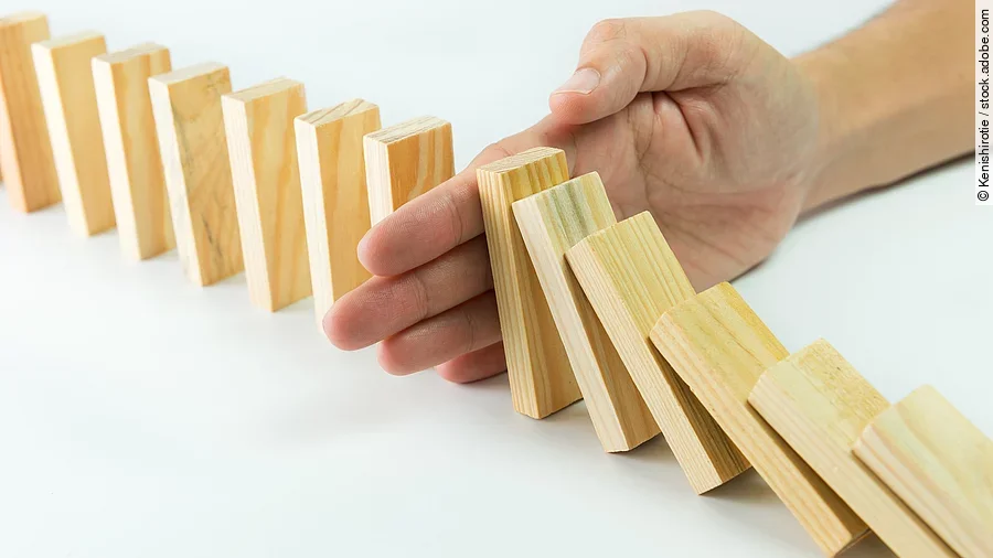 Hölzerne Dominosteine sind in einer Reihe aufgebaut. Einige davon sind bereits umgefallen, eine Hand stoppt den Dominoeffekt.