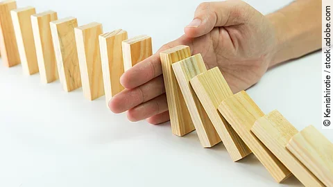 Stop Hölzerne Dominosteine sind in einer Reihe aufgebaut. Einige davon sind bereits umgefallen, eine Hand stoppt den Dominoeffekt.
