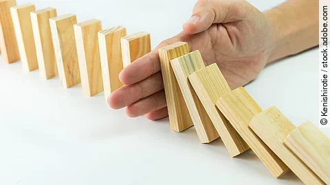 Hölzerne Dominosteine sind in einer Reihe aufgebaut. Einige davon sind bereits umgefallen, eine Hand stoppt den Dominoeffekt.