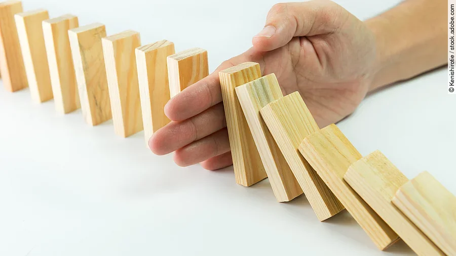 Stop Hölzerne Dominosteine sind in einer Reihe aufgebaut. Einige davon sind bereits umgefallen, eine Hand stoppt den Dominoeffekt.