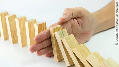 Hölzerne Dominosteine sind in einer Reihe aufgebaut. Einige davon sind bereits umgefallen, eine Hand stoppt den Dominoeffekt.
