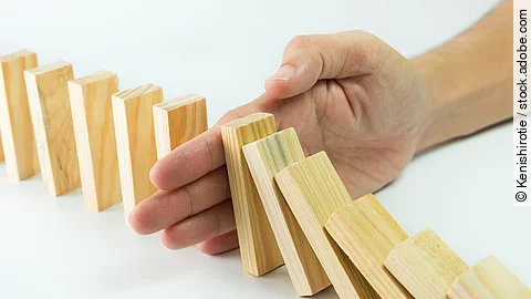 Stop Hölzerne Dominosteine sind in einer Reihe aufgebaut. Einige davon sind bereits umgefallen, eine Hand stoppt den Dominoeffekt.