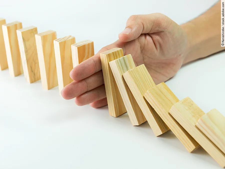 Hölzerne Dominosteine sind in einer Reihe aufgebaut. Einige davon sind bereits umgefallen, eine Hand stoppt den Dominoeffekt.
