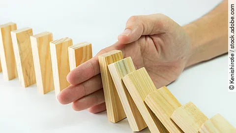 Stop Hölzerne Dominosteine sind in einer Reihe aufgebaut. Einige davon sind bereits umgefallen, eine Hand stoppt den Dominoeffekt.