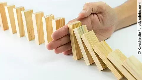 Hölzerne Dominosteine sind in einer Reihe aufgebaut. Einige davon sind bereits umgefallen, eine Hand stoppt den Dominoeffekt.