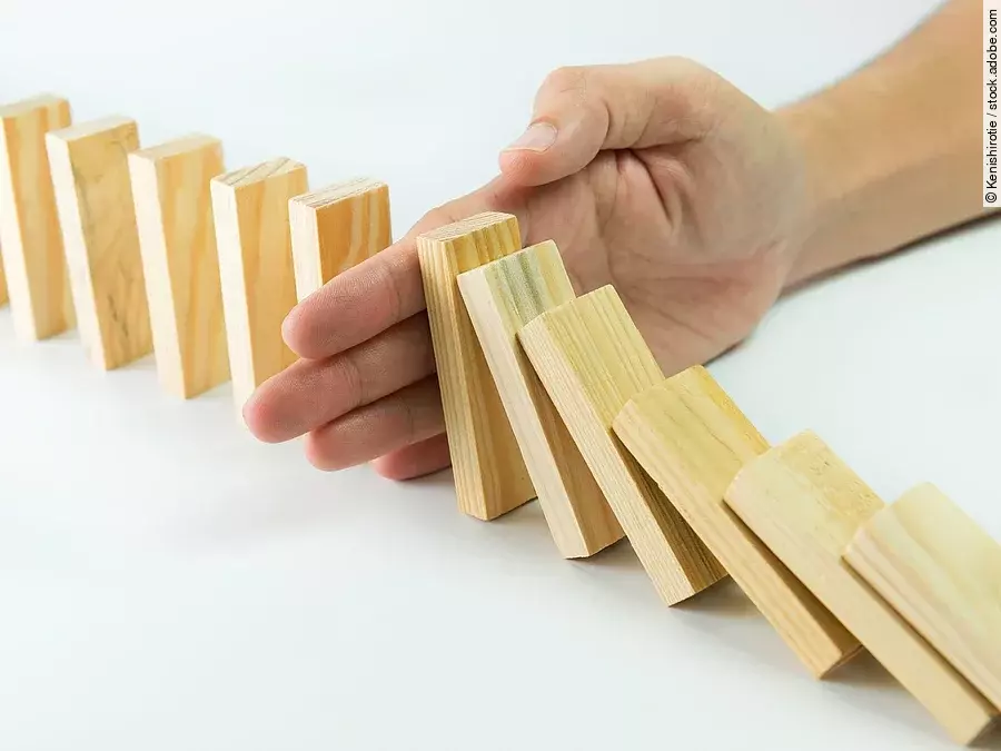 Stop Hölzerne Dominosteine sind in einer Reihe aufgebaut. Einige davon sind bereits umgefallen, eine Hand stoppt den Dominoeffekt.