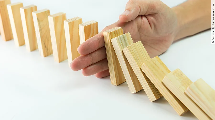 Hölzerne Dominosteine sind in einer Reihe aufgebaut. Einige davon sind bereits umgefallen, eine Hand stoppt den Dominoeffekt.