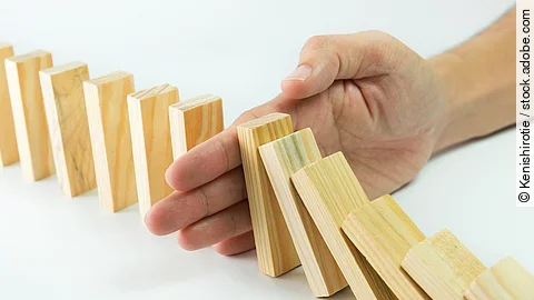 Stop Hölzerne Dominosteine sind in einer Reihe aufgebaut. Einige davon sind bereits umgefallen, eine Hand stoppt den Dominoeffekt.