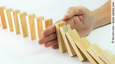 Hölzerne Dominosteine sind in einer Reihe aufgebaut. Einige davon sind bereits umgefallen, eine Hand stoppt den Dominoeffekt.