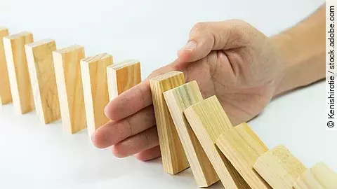 Stop Hölzerne Dominosteine sind in einer Reihe aufgebaut. Einige davon sind bereits umgefallen, eine Hand stoppt den Dominoeffekt.