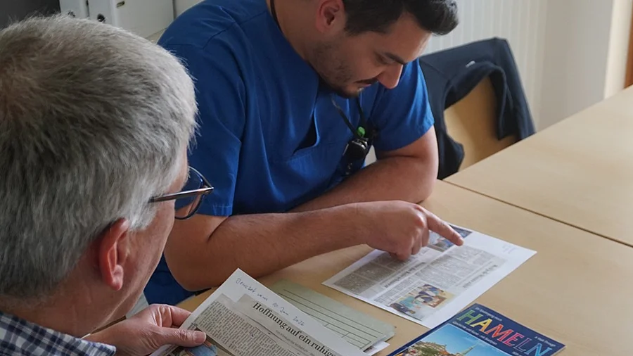 Sprachcafé am Sana Klinikum Hameln-Pyrmont Links ein älterer Mann der einen Zeitungsausschnitt hält, rechts ein Mann in blauem Kasack, der einen Zeitungsausschnitt liest