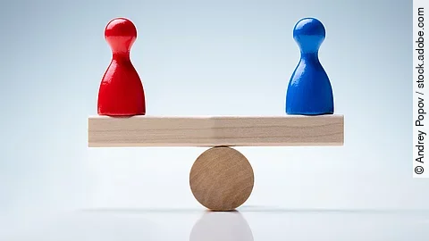 Auf einer holzähnlichen Waage stehen eine rote und blaue Figur, die das Gleichgewicht halten. 