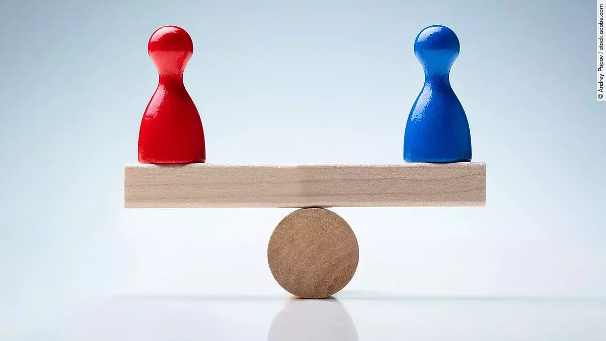 Auf einer holzähnlichen Waage stehen eine rote und blaue Figur, die das Gleichgewicht halten. 