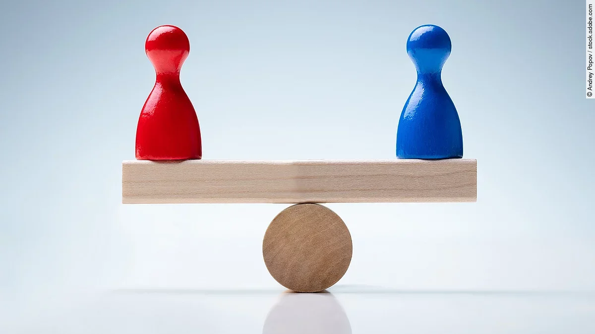 Gleichheit Auf einer holzähnlichen Waage stehen eine rote und blaue Figur, die das Gleichgewicht halten.
