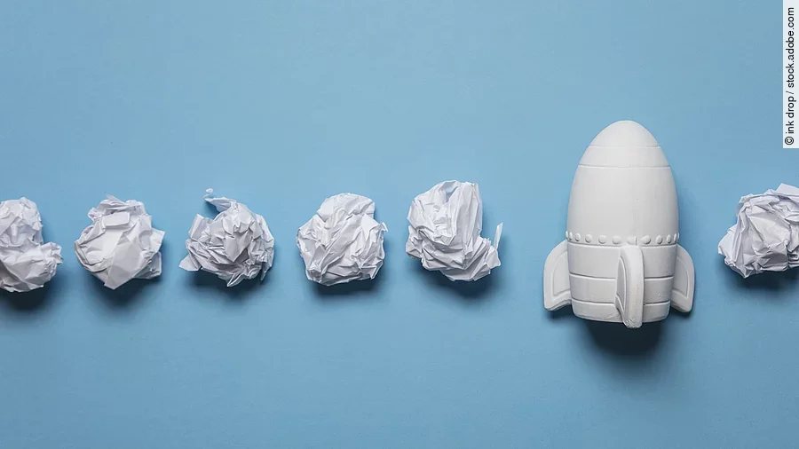 Start-up In einer Reihe liegen zusammengeknüllte Blätter Papier. Rechts in der Reihe liegt statt einem Papier eine kleine weiße Rakete.