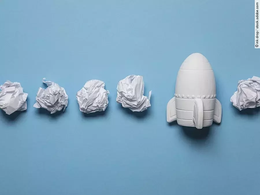 Start-up In einer Reihe liegen zusammengeknüllte Blätter Papier. Rechts in der Reihe liegt statt einem Papier eine kleine weiße Rakete.
