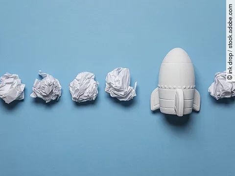 In einer Reihe liegen zusammengeknüllte Blätter Papier. Rechts in der Reihe liegt statt einem Papier eine kleine weiße Rakete.