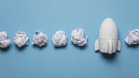 Start-up In einer Reihe liegen zusammengeknüllte Blätter Papier. Rechts in der Reihe liegt statt einem Papier eine kleine weiße Rakete.