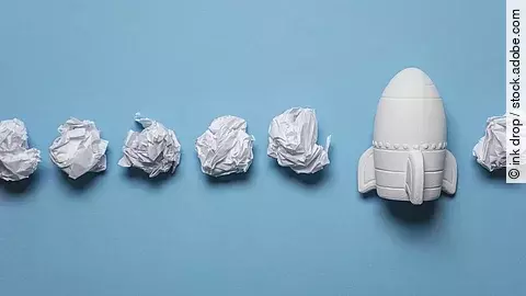 In einer Reihe liegen zusammengeknüllte Blätter Papier. Rechts in der Reihe liegt statt einem Papier eine kleine weiße Rakete.