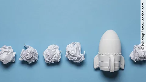 Start-up In einer Reihe liegen zusammengeknüllte Blätter Papier. Rechts in der Reihe liegt statt einem Papier eine kleine weiße Rakete.