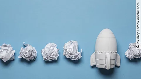 In einer Reihe liegen zusammengeknüllte Blätter Papier. Rechts in der Reihe liegt statt einem Papier eine kleine weiße Rakete.