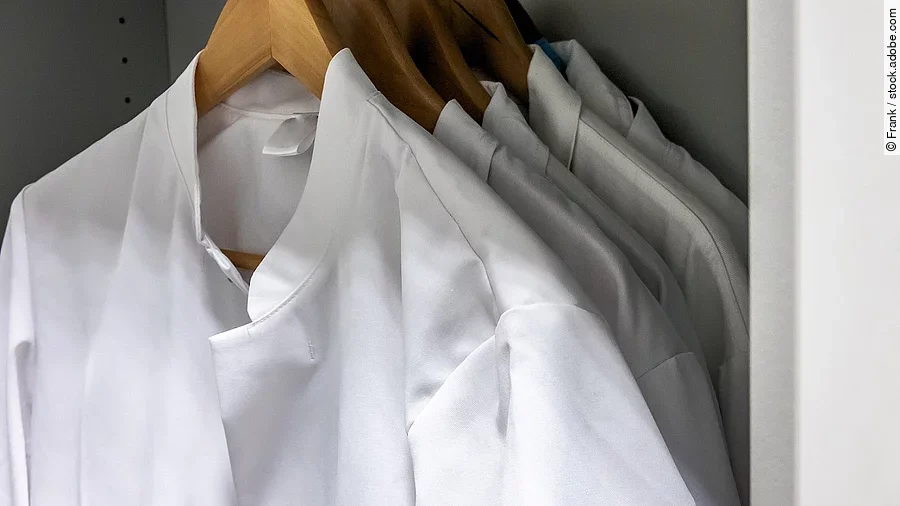 Mehrere Arztkittel hängen auf braunen Kleiderbügeln im Schrank.