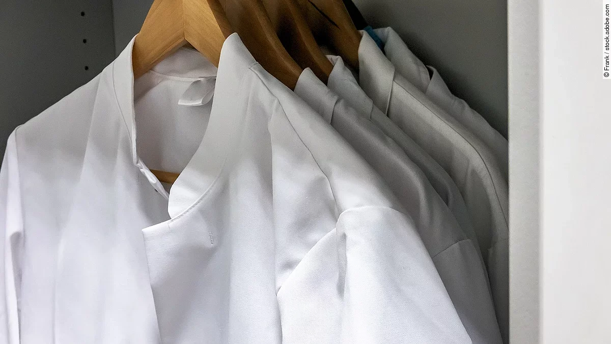 Mehrere Arztkittel hängen auf braunen Kleiderbügeln im Schrank.