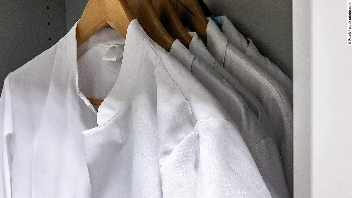 Mehrere Arztkittel hängen auf braunen Kleiderbügeln im Schrank.