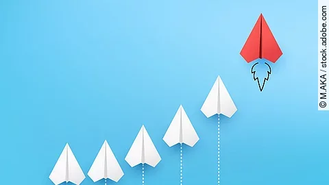 Sechs Papierflieger, die kurvenähnlich angeordnet sind vor blauem Hintergrund. Der rechte und höchste Papierflieger ist rot.