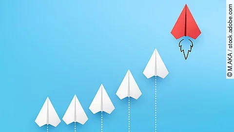 Sechs Papierflieger, die kurvenähnlich angeordnet sind vor blauem Hintergrund. Der rechte und höchste Papierflieger ist rot.