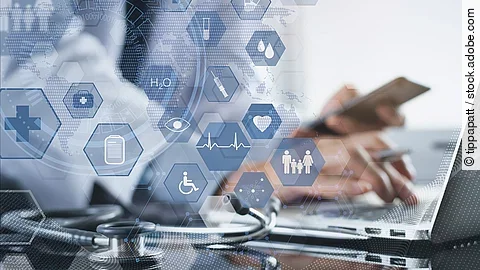 Ein Mann sitzt an seinem Laptop. Daneben liegt ein Stethoskop. Im Vordergrund erscheint eine holografische Darstellung aus vielen einzelnen Icons und Symbolen, die ein digitales Netzwerk aus Daten verkörpern sollen.