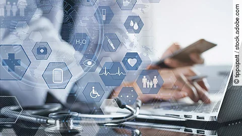 Cloud Ein Mann sitzt an seinem Laptop. Daneben liegt ein Stethoskop. Im Vordergrund erscheint eine holografische Darstellung aus vielen einzelnen Icons und Symbolen, die ein digitales Netzwerk aus Daten verkörpern sollen.