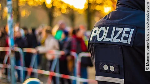 Demonstration im Vordergrund ist der Rücken eines Polizisten in Uniform. Im Hintergrund stehen mehrere Personen vor Absperrband.