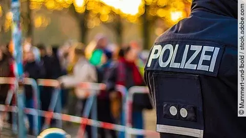 im Vordergrund ist der Rücken eines Polizisten in Uniform. Im Hintergrund stehen mehrere Personen vor Absperrband.