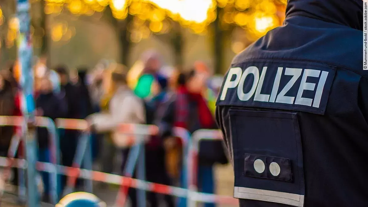 Demonstration im Vordergrund ist der Rücken eines Polizisten in Uniform. Im Hintergrund stehen mehrere Personen vor Absperrband.