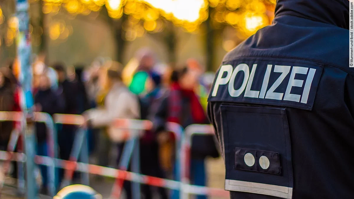 Demonstration im Vordergrund ist der Rücken eines Polizisten in Uniform. Im Hintergrund stehen mehrere Personen vor Absperrband.