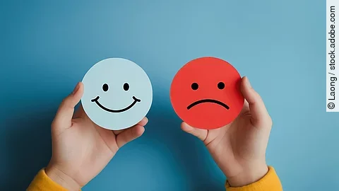 Zwei Hände halten ein fröhliches und ein trauriges Gesicht gemalt auf verschiedenfarbigen Papierausschnitten