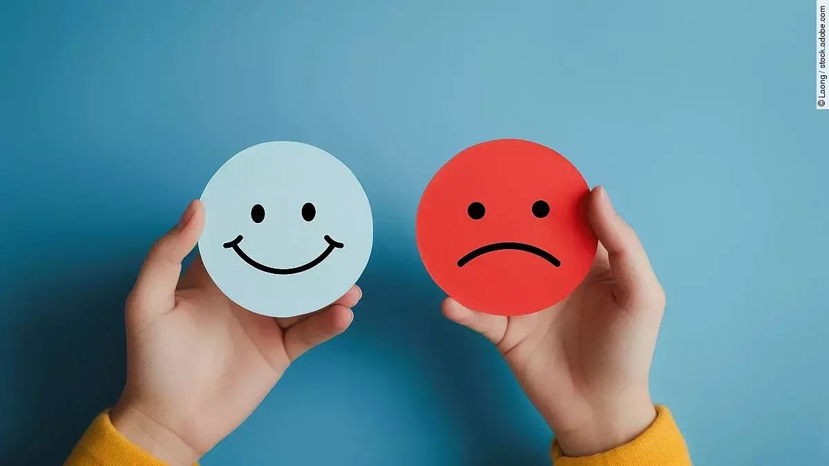 Zwei Hände halten ein fröhliches und ein trauriges Gesicht gemalt auf verschiedenfarbigen Papierausschnitten