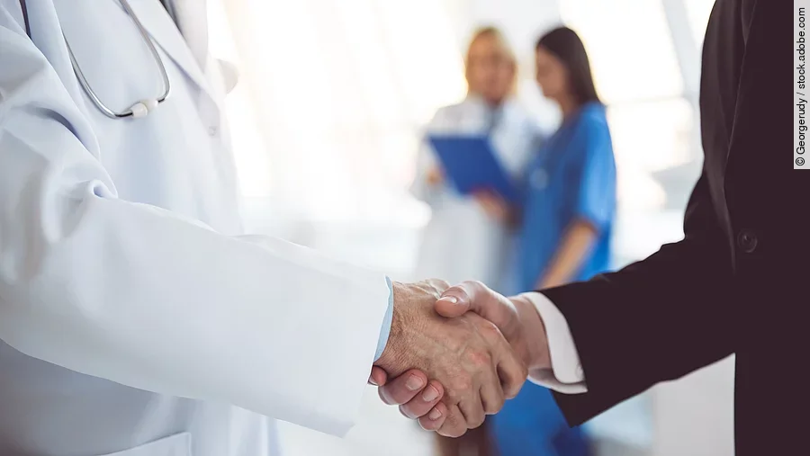 Ein Arzt und ein Geschäftsmann schütteln Hände. Im Hintergrund sind zwei Krankenhausmitarbeiterinnen verschwommen zu sehen, eine Ärztin und eine Pflegerin.