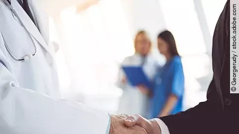 Ein Arzt und ein Geschäftsmann schütteln Hände. Im Hintergrund sind zwei Krankenhausmitarbeiterinnen verschwommen zu sehen, eine Ärztin und eine Pflegerin.