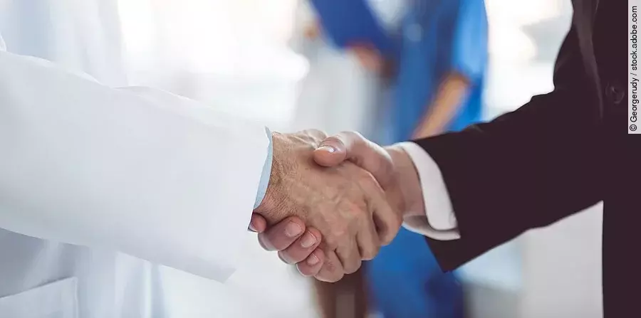 Ein Arzt und ein Geschäftsmann schütteln Hände. Im Hintergrund sind zwei Krankenhausmitarbeiterinnen verschwommen zu sehen, eine Ärztin und eine Pflegerin.