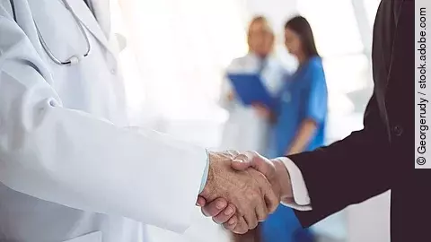Ein Arzt und ein Geschäftsmann schütteln Hände. Im Hintergrund sind zwei Krankenhausmitarbeiterinnen verschwommen zu sehen, eine Ärztin und eine Pflegerin.