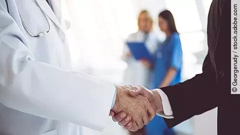 Ein Arzt und ein Geschäftsmann schütteln Hände. Im Hintergrund sind zwei Krankenhausmitarbeiterinnen verschwommen zu sehen, eine Ärztin und eine Pflegerin.