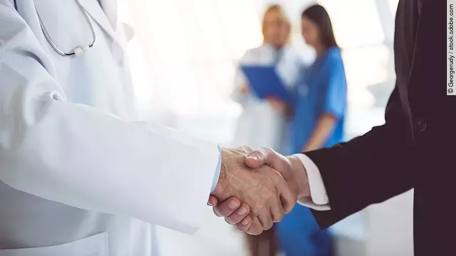 Ein Arzt und ein Geschäftsmann schütteln Hände. Im Hintergrund sind zwei Krankenhausmitarbeiterinnen verschwommen zu sehen, eine Ärztin und eine Pflegerin.