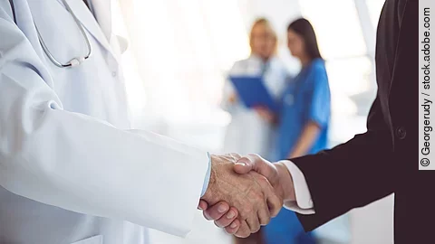 Händeschütteln Ein Arzt und ein Geschäftsmann schütteln Hände. Im Hintergrund sind zwei Krankenhausmitarbeiterinnen verschwommen zu sehen, eine Ärztin und eine Pflegerin.
