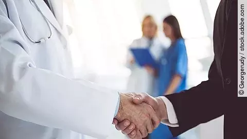 Ein Arzt und ein Geschäftsmann schütteln Hände. Im Hintergrund sind zwei Krankenhausmitarbeiterinnen verschwommen zu sehen, eine Ärztin und eine Pflegerin.